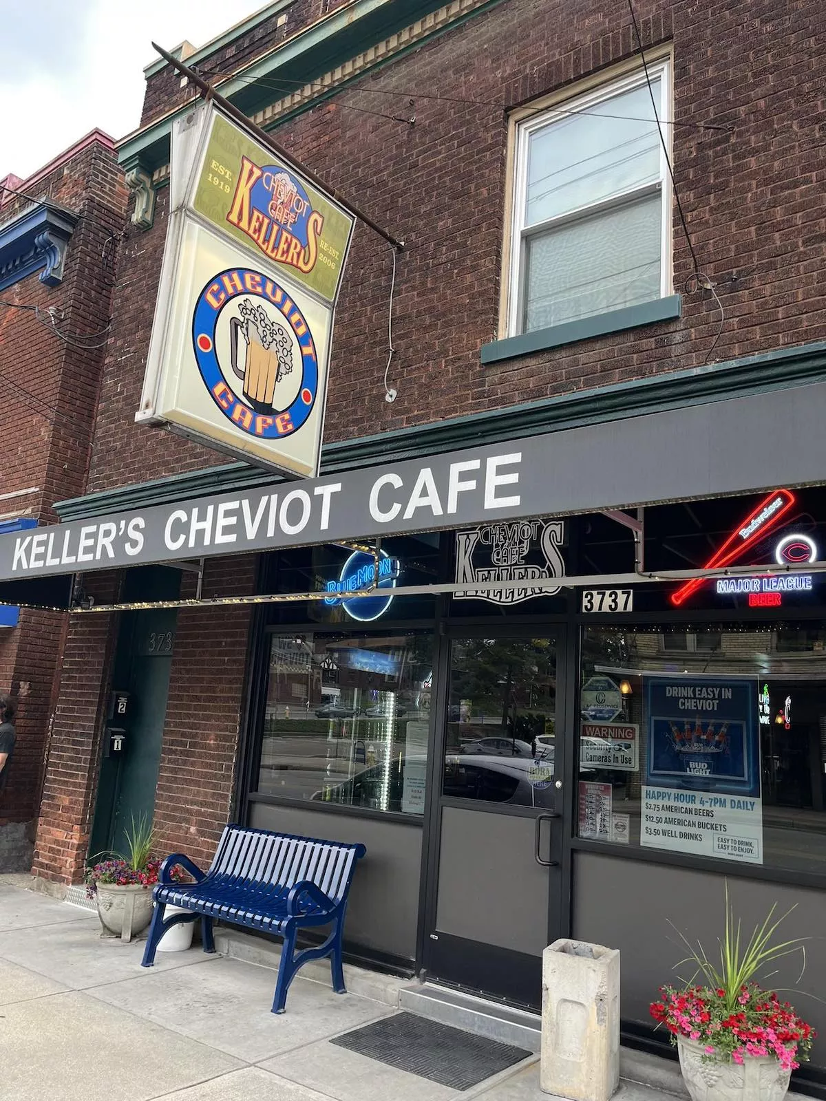 Keller's Cheviot Cafe storefront on Glenmore Avenue, with its vintage hanging sign, blue bench, and flower planters